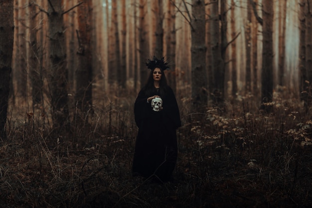 terrible witch holds the skull of a dead man in her hands in a dark forest