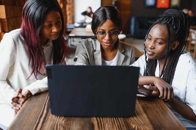 Three African American girls students sitting at the table in cafe studying up for test or making homework together they are using laptop and digital tablet