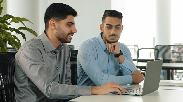 Two Indian employees sitting in modern office using laptop young colleagues discussing project Arab serious millennial male businessmen satisfied with result of negotiations concluded contract