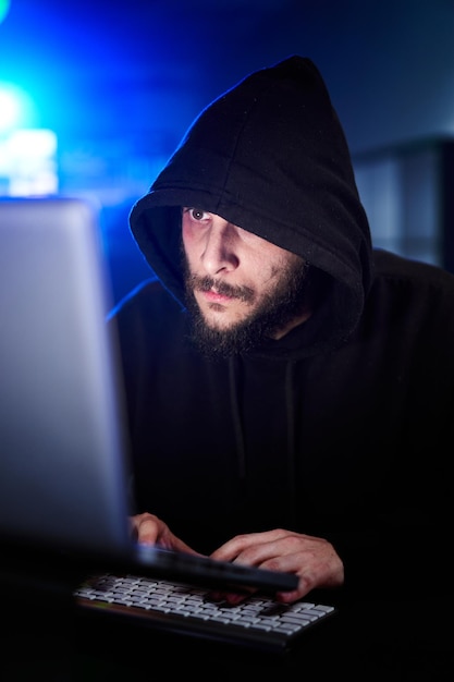 vertical portrait of mysterious hooded hacker sitting at desk and breaking into government data serv