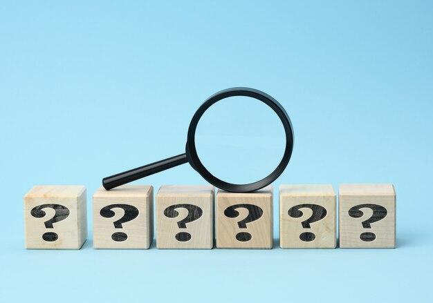Wooden cubes with question marks and a magnifying glass on a blue background. The concept of finding answers to unknown questions, solving a problem. Find information