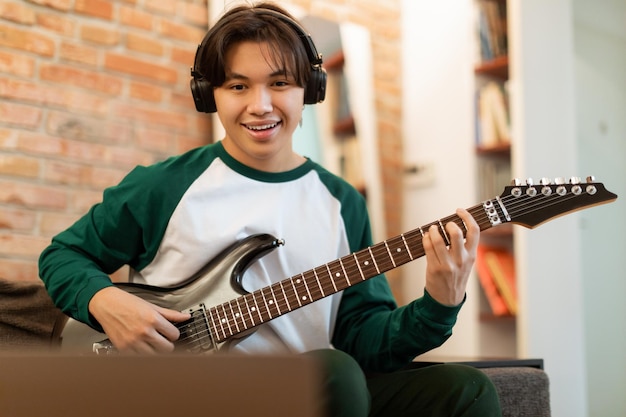 young asian guy practicing guitar chords looking at laptop indoor