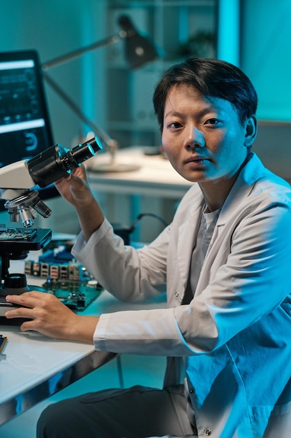 Young chinese woman in whitecoat looking at camera while working with microscope