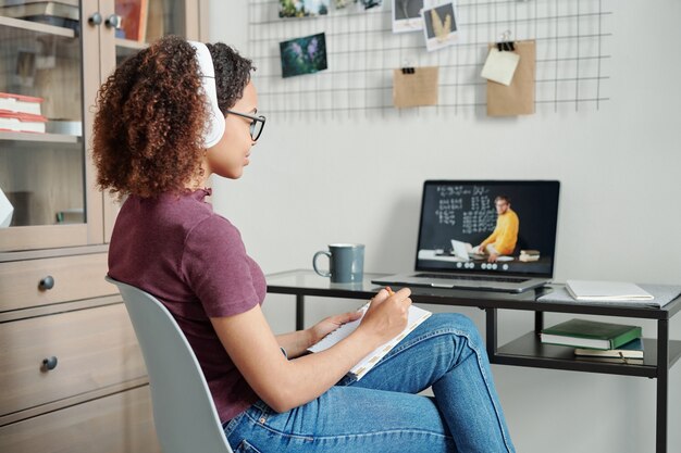 Young diligent female student in casualwear and headphones making notes in front of laptop while listening to teacher during online lesson