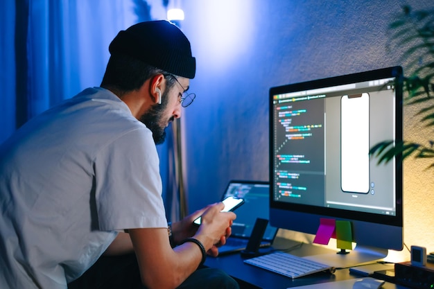 Young man mobile developer writes program code on a computer and check app in smartphone programmer work in home office