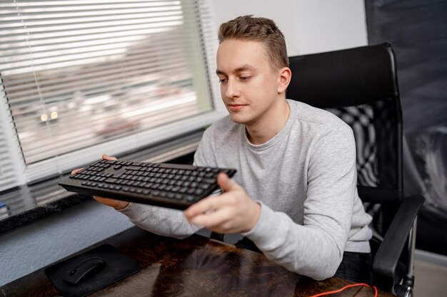 Young man sits at table and holds computer keyboard in his hands