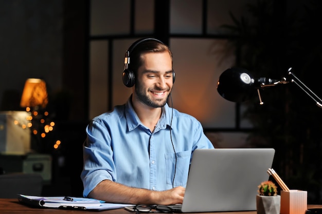 Young man using laptop and listening to music in office at night