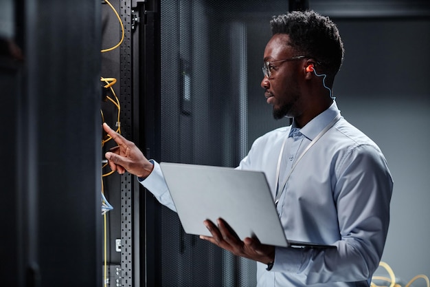 Young network engineer working with servers in data center and holding laptop