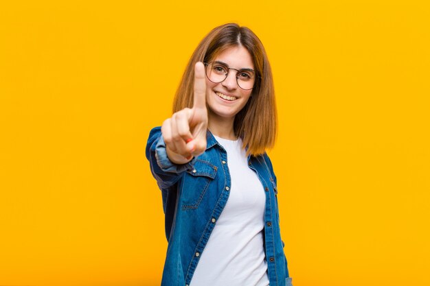 Young pretty woman smiling proudly and confidently making number one pose triumphantly, feeling like a leader over yellow wall