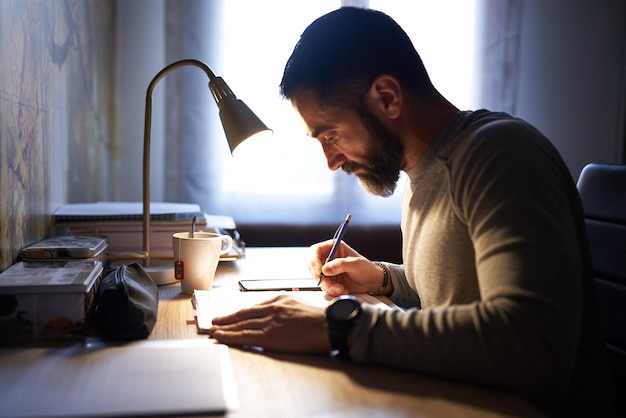 Young white man with a neat beard studying and looking at the phone at his desk