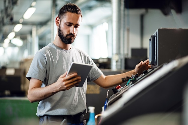 Young worker operating a CNC machine while using digital tablet in industrial facility