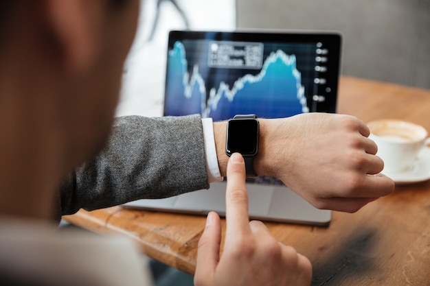 Cropped image of businessman sitting by the table in cafe and analyzing indicators on laptop computer while using wristwatch