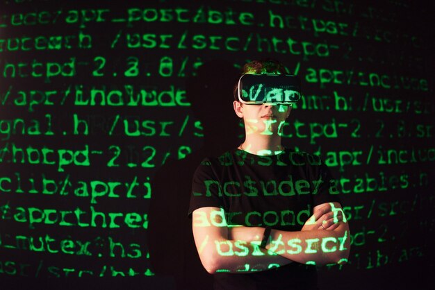 Double exposure of a caucasian man and Virtual reality VR headset is presumably a gamer or a hacker cracking the code into a secure network or server, with lines of code in green