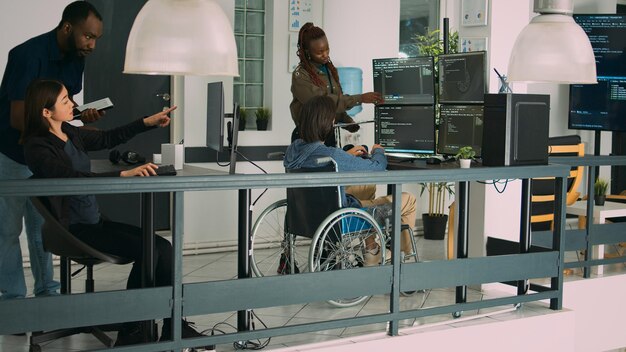 Asian coder with physical disability working with african american developer to create security firewall on terminasl window. IT engineer in wheelchair programming html script code.