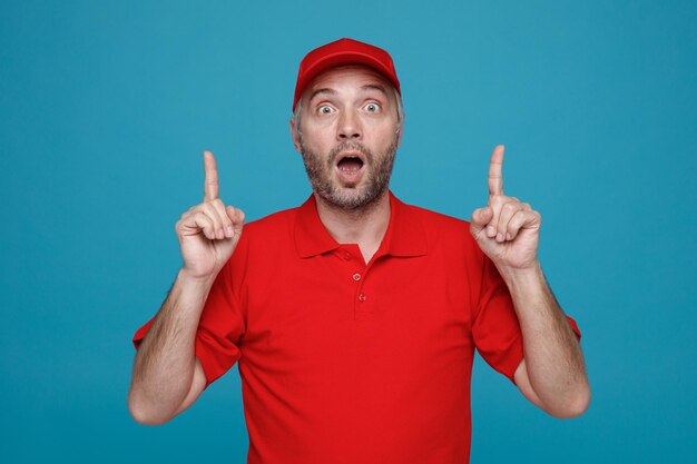 delivery man employee in red cap blank tshirt uniform looking at camera amazed and surprised pointing with index fingers up with both hands standing over blue background