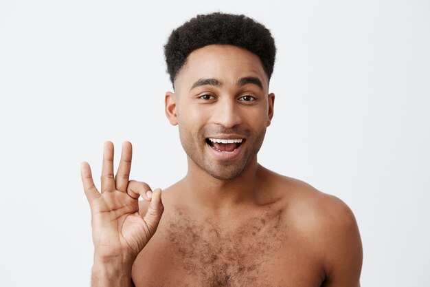 Ça va. Ne t'inquiète pas. Gros plan de gai jeune homme séduisant à la peau sombre avec une coiffure afro avec torse nu montrant signe OK avec la main, un clin de œil, regardant à huis clos avec une expression affectueuse.