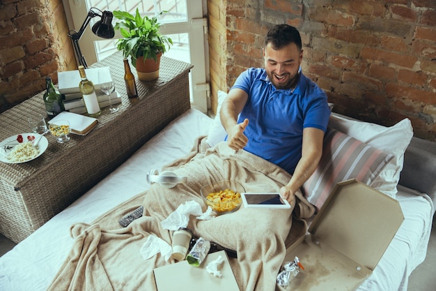 Acclamations enthousiastes de l'équipe sportive, pouce vers le haut. Homme paresseux vivant dans son lit entouré de désordre. Pas besoin de sortir pour être heureux. Utiliser des gadgets, regarder des films et des séries, émotif. Fast food.