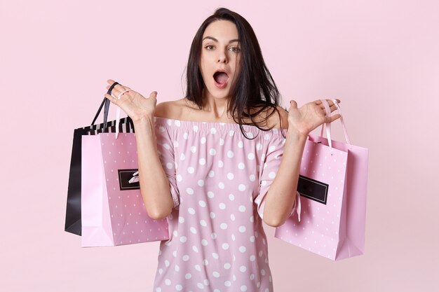 Accro du shopping femme surprise vêtue d'une robe élégante, tient des sacs à deux mains, oublie d'acheter quelque chose, se sent choquée de voir de grosses remises au magasin, isolé sur un mur rose