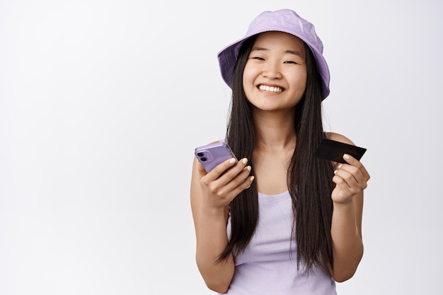 Achats en ligne Belle fille asiatique tenant une carte de crédit et un smartphone souriant heureux à la caméra faisant un bon de commande dans l'application fond blanc