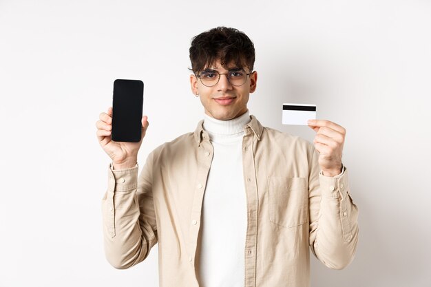 Achats en ligne jeune homme moderne montrant une carte de crédit en plastique et un écran de smartphone vide démontrant...