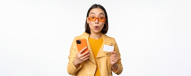 Achats en ligne Modèle féminin asiatique élégant à lunettes de soleil tenant une carte de crédit et un téléphone portable souriant heureux debout sur fond blanc