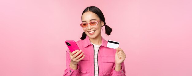 Achats en ligne Smiling asian girl shopper holding smartphone et carte de crédit payant dans l'application mobile debout sur fond rose