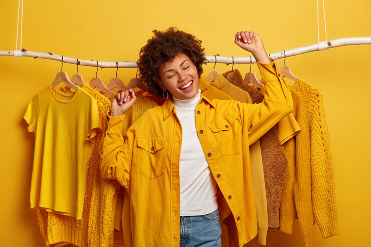Photo gratuit l'acheteur de vêtements bouclés insouciants danse avec bonheur, lève les bras, achète des vêtements jaunes de la nouvelle collection, se réjouissant d'une journée de magasinage réussie, étant de bonne humeur, danse contre des tenues sur un support