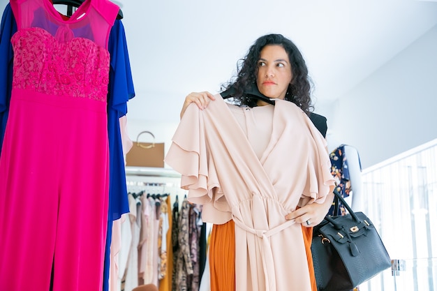 Acheteuse sérieuse tenant un cintre avec un chiffon, appliquant une robe sur elle-même, regardant ailleurs. Vue de face. Magasin de mode ou concept de vente au détail