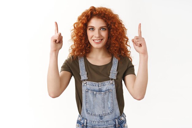 Actrice rousse ambitieuse rêvant de devenir le top pointant les doigts vers le haut avec une expression confiante souriante avec un sourire à pleines dents blanc déterminé à réussir debout fond blanc