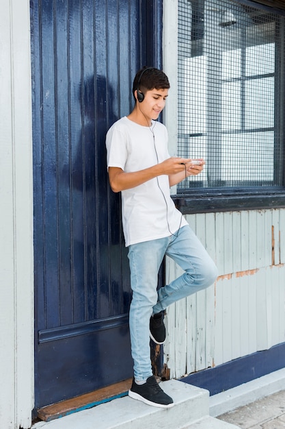 Adolescent heureux debout à la porte bleue fermée à l&#39;aide de téléphone portable avec un casque sur sa tête