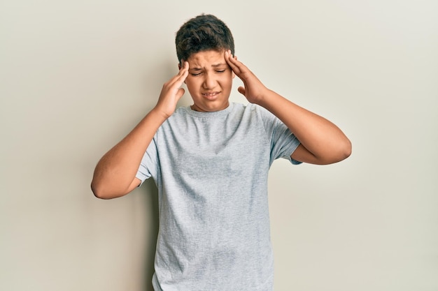 Adolescent hispanique portant un t-shirt gris décontracté avec la main sur la tête pour la douleur dans la tête à cause du stress. souffrant de migraine.