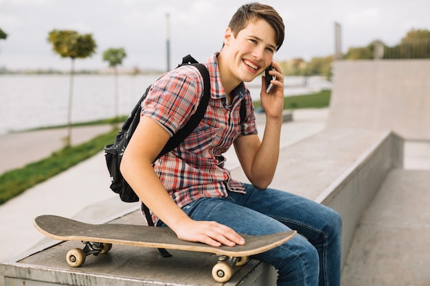 Photo gratuite adolescent joyeux parler au téléphone près de planche à roulettes