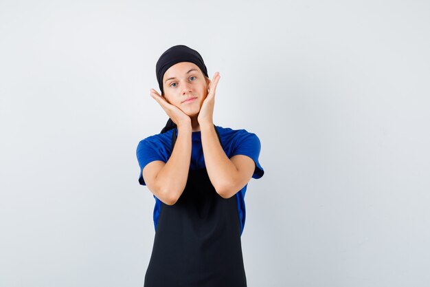 Un adolescent masculin cuisinier en t-shirt, un tablier gardant les mains sur les joues et l'air confiant, vue de face.