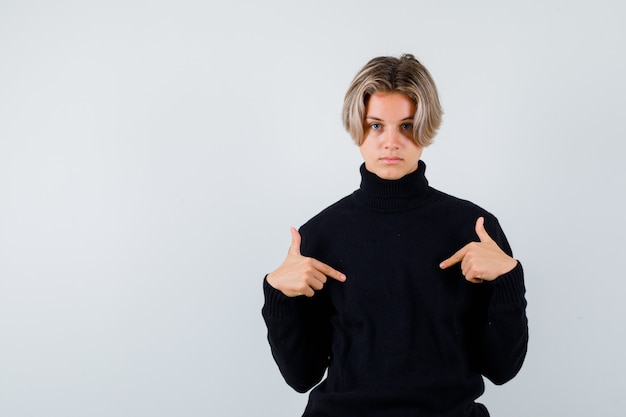 Adolescent mignon pointant sur lui-même dans un pull à col roulé noir et l'air perplexe, vue de face.