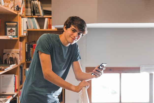 Adolescent souriant avec smartphone dans la bibliothèque