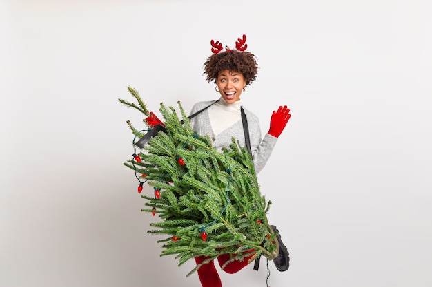 Photo gratuite adolescente afro-américaine assez joyeuse soulève des danses de main et de jambe sans soucis tient le sapin comme si un instrument de musique prétend être un guitariste professionnel porte un costume de noël s'amuse à la fête