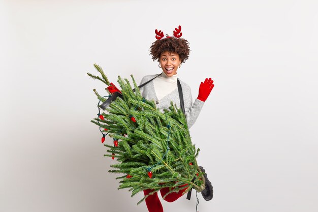 Adolescente afro-américaine assez joyeuse soulève des danses de main et de jambe sans soucis tient le sapin comme si un instrument de musique prétend être un guitariste professionnel porte un costume de Noël s'amuse à la fête
