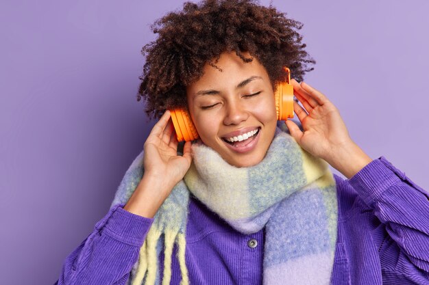 Une adolescente afro-américaine optimiste insouciante sourit largement garde les mains sur des écouteurs stéréo écoute de la musique garde les yeux fermés porte une écharpe chaude autour du cou.