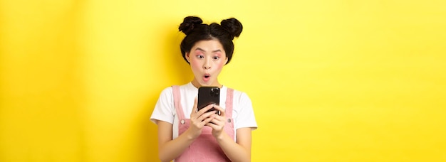 Une adolescente asiatique a l'air excitée à l'écran du smartphone lisant les nouvelles sur le téléphone debout sur un motif jaune