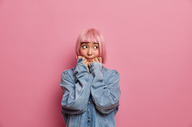 Photo gratuite adolescente asiatique aux cheveux roses, garde les mains sous le menton et détourne le regard avec une expression réfléchie, vêtue d'une veste en jean surdimensionnée, essaie de se souvenir de quelque chose, imagine comment résoudre le problème