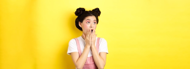 Une adolescente asiatique choquée avec du maquillage haletant et couvrant la bouche regardant à gauche le logo étonné debout