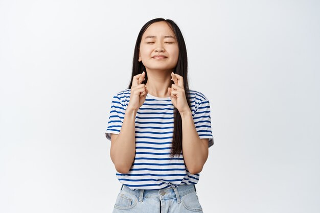 Une adolescente asiatique pleine d'espoir fait un vœu, ferme les yeux et croise les doigts pour la bonne chance, prie, anticipe que qch arrive, debout sur blanc.
