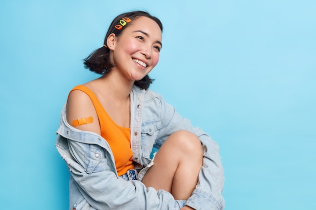 Photo gratuite une adolescente asiatique positive avec deux queues de cheval regarde joyeusement de côté montre une épaule plâtrée heureuse après avoir été vaccinée se sent protégée porte une veste en jean élégante isolée sur un mur bleu