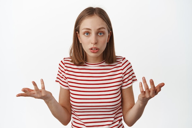 Une Adolescente Blonde Confuse Haussant Les épaules, Levant Les Mains Et Ayant L'air Perplexe, Ne Peut Pas Comprendre Ce Qui S'est Passé, Posant Des Questions, Regardant Sans Aucune Idée Devant, Debout Sur Un Mur Blanc.