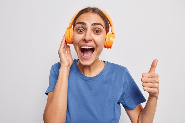 Une adolescente brune joyeuse s'exclame bruyamment montre un geste correct garde le pouce vers le haut recommande sa liste de lecture porte un casque stéréo orange t-shirt bleu décontracté isolé sur fond de studio blanc