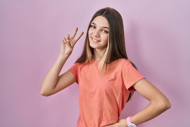 Adolescente debout sur fond rose souriant regardant la caméra montrant les doigts faisant le signe de la victoire numéro deux