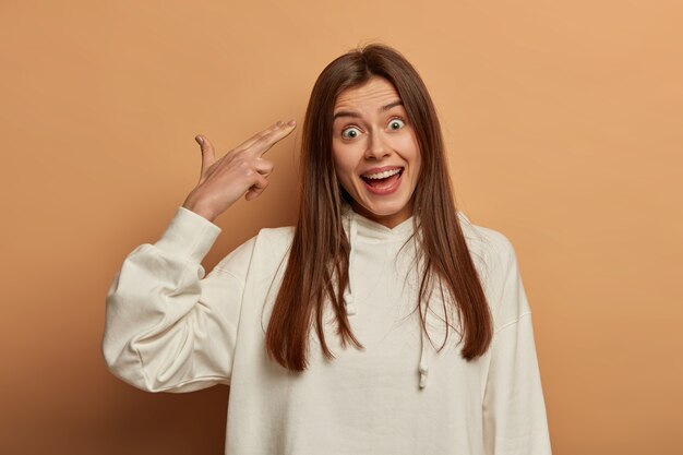 Une adolescente drôle se trompe, tire dans le temple, fait semblant de se suicider, a une expression joyeuse, porte un sweat-shirt blanc