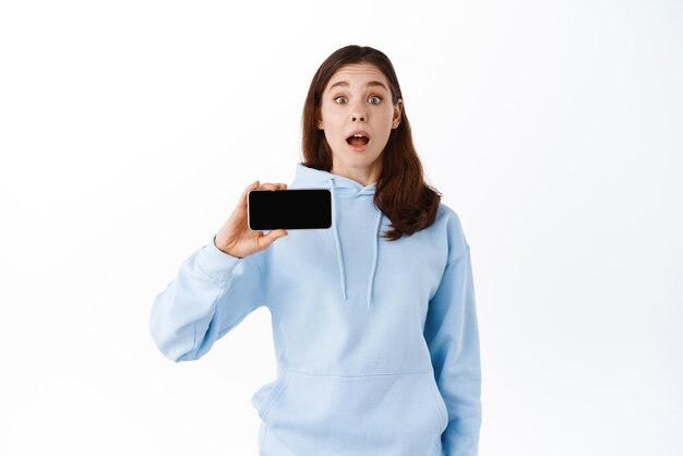 Adolescente excitée montrant une application ou un jeu vidéo sur un écran de téléphone portable vide démontre votre promo sur l'écran du smartphone debout sur fond blanc