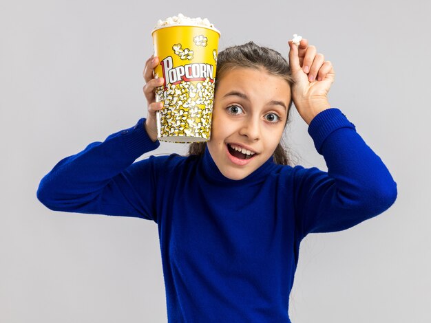 Adolescente excitée tenant un seau de pop-corn et un morceau de pop-corn touchant la tête avec un seau de pop-corn et une main isolée sur un mur blanc