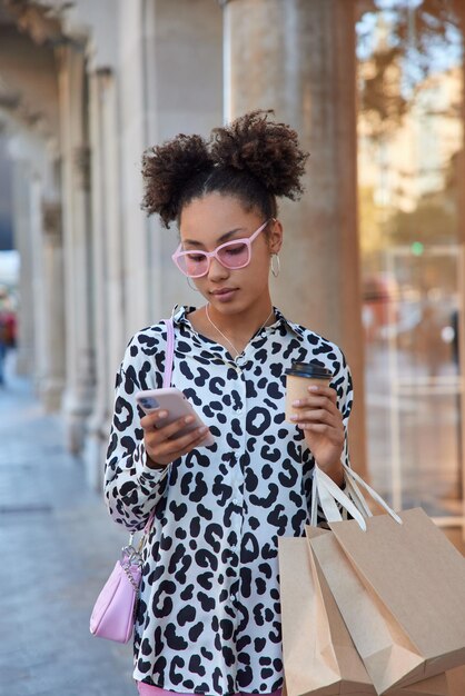 Une adolescente frisée à la mode porte des lunettes de soleil roses à la mode et un chemisier boit du café à emporter utilise des chèques cellulaires la notification porte des sacs en papier pose dans la rue de la ville profite du week-end